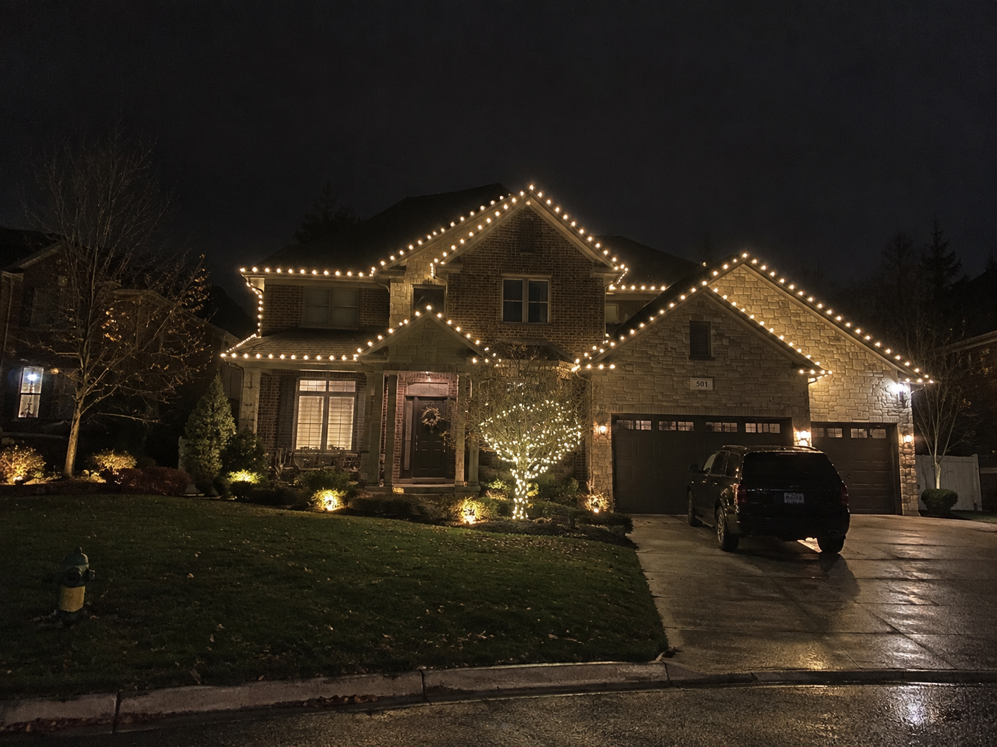 Brick and stone home with warm white roofline lights and a lit tree at night